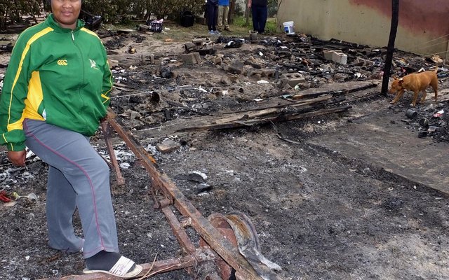 Abrien Fortuin staan op die plek waar haar karavaan gestaan het voordat dit deur die brand verwoes is, met die huise waarvan die vensters uitgeruk is in die agtergrond.